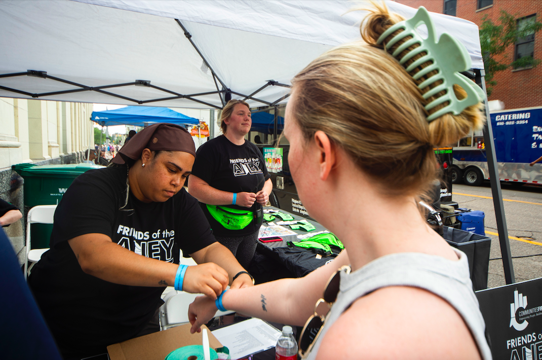 Kristin Stevenson, president of local nonprofit Friends of the Alley, left, places a wristband onto the wrist of an attendee as hundreds of people gather in downtown Flint's Buckham Alley for Alley Fest on Saturday, July 8, 2023. (Katy Kildee | Flintside)