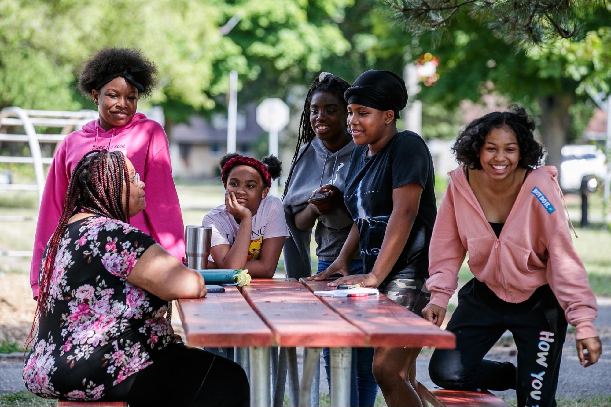 Honny Williams sits on a park bench while talking to neighborhood kids (pictured from left to right; Terynn Henry, Sakara Johnsonn, Nene, Miesia Leroy, Nikiyah Johnson) in Sarvis Park, Flint on July 26, 2022. (Jenifer Veloso | Flintside.com)