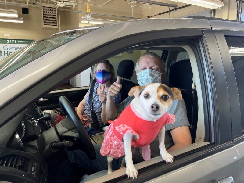 A couple gives a thumbs up from their vehicle at a previous Genesee Health Plan drive-thru vaccine event as their cute pet looks on.