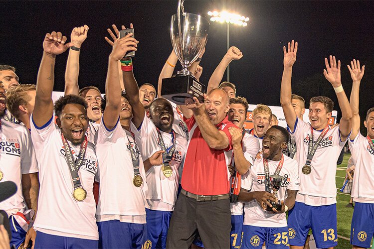 Costa Papista holds up the Bucks' hard-earned USL League Two Championship trophy as Bucks team members celebrate the victory last year.